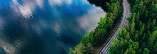 Cars driving on forested road next to lake that reflects the sky