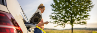 Woman scrolling on smart phone while charging electric vehicle next to tree